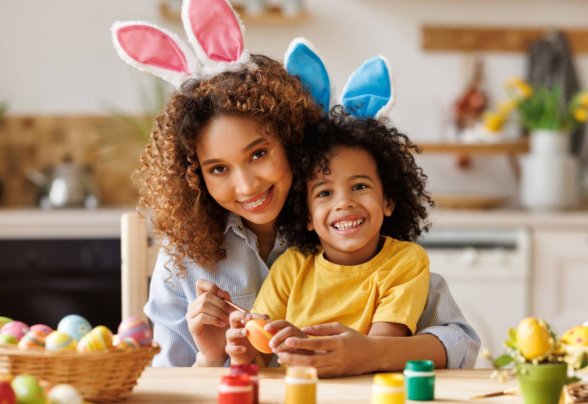 Happy african american family: mother teaching happy little kid soon to decorate Easter eggs while sitting in kitchen